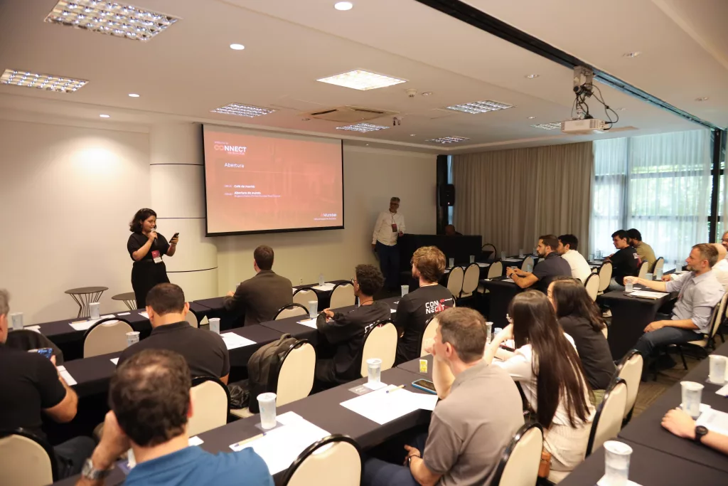 Mulher palestrando em evento corporativo com tela de projeção e público atento.
